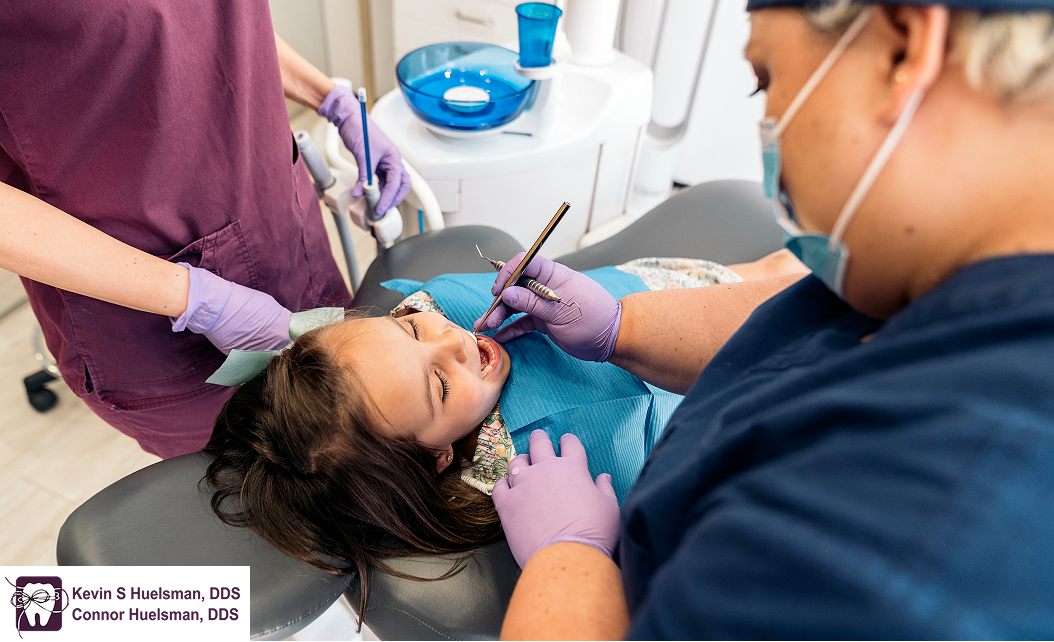 Child having a dental checkup by dentist while sitting beside the assistant wearing maroon uniform